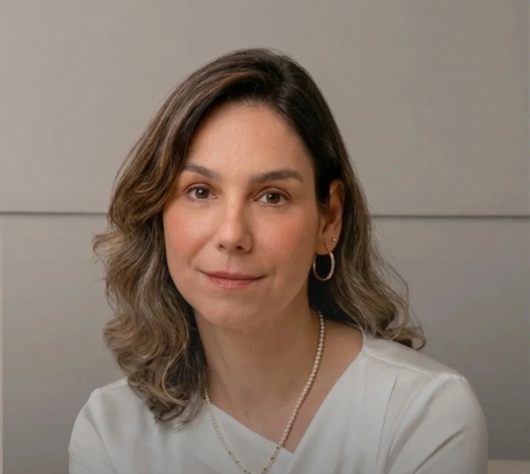 Dra. Eveline Saad, de blusa branca e colar de pérolas, diante do painel cinza do estúdio